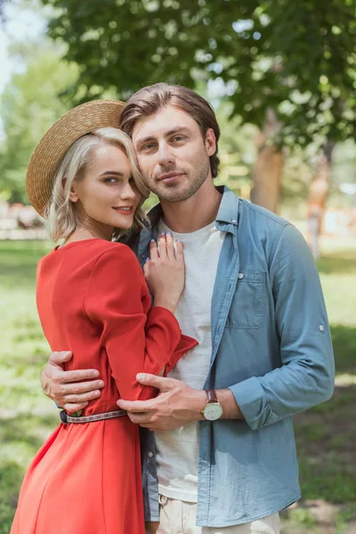 Souriant petite amie et copain étreignant dans le parc et regardant la caméra — Photo de stock