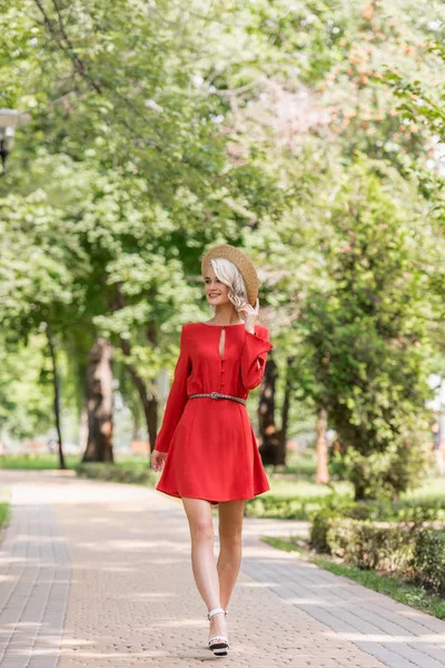 Lächelndes schönes Mädchen in rotem Kleid und Strohhut, das im Park spaziert und wegschaut — Stockfoto