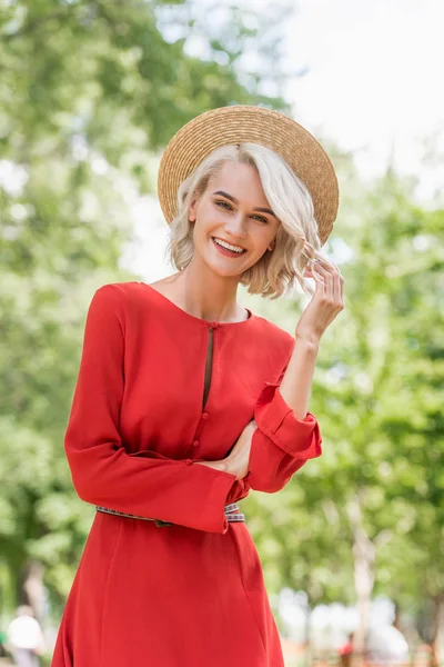 Souriant attrayant fille en robe rouge et chapeau de paille regardant caméra dans le parc — Photo de stock