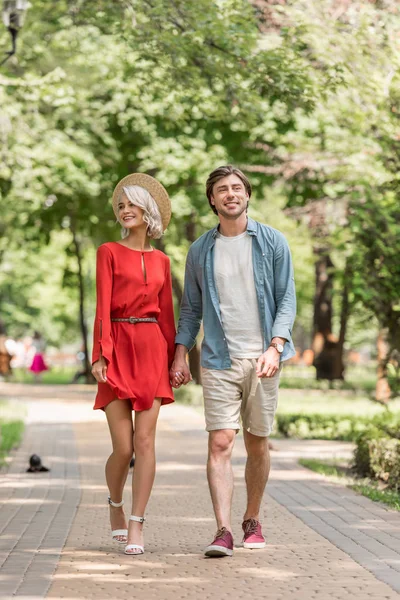Novia sonriente y novio tomados de la mano y caminando juntos en el parque - foto de stock