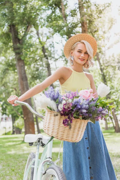 Belle fille blonde tenant vélo avec panier de fleurs dans le parc — Photo de stock