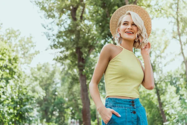 Low angle view of stylish attractive blonde girl in straw hat looking away in park — Stock Photo