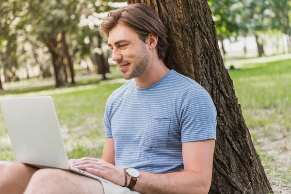 Sorrindo bonito homem sentado perto da árvore no parque e usando laptop — Fotografia de Stock