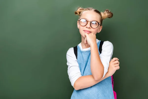 Pensive schoolchild in glasses looking up near blackboard — Stock Photo