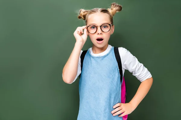 Shocked schoolchild touching glasses and looking at camera near blackboard — Stock Photo