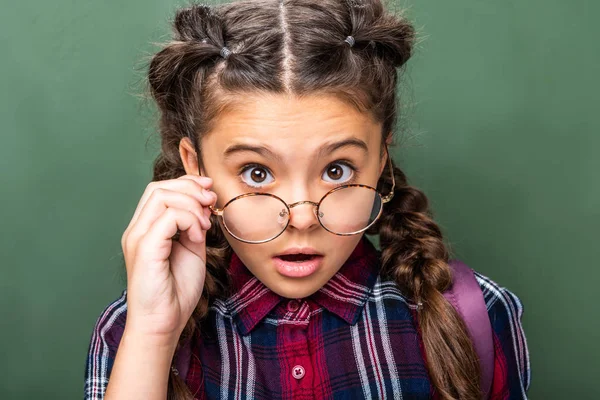 Surprised schoolchild looking at camera above glasses near blackboard — Stock Photo