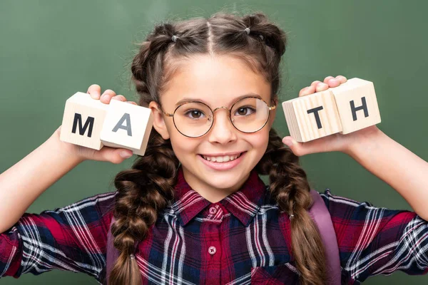 Retrato de criança em idade escolar segurando cubos de madeira com palavra matemática perto de quadro negro — Fotografia de Stock