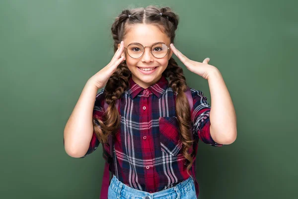 Smiling schoolchild touching head and looking at camera near blackboard — Stock Photo