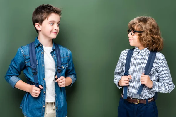 Schoolboys with backpacks looking at each other near blackboard — Stock Photo