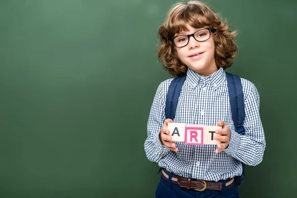 Schoolboy holding wooden cubes with word art near blackboard — Stock Photo