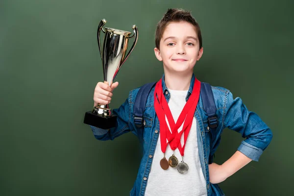 Schoolboy with medals holding winner cup near blackboard — Stock Photo
