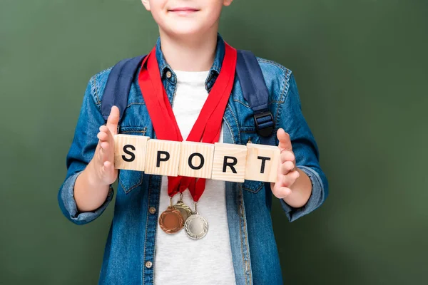 Image recadrée d'écolier avec des médailles tenant des cubes en bois avec mot sport près du tableau noir — Photo de stock