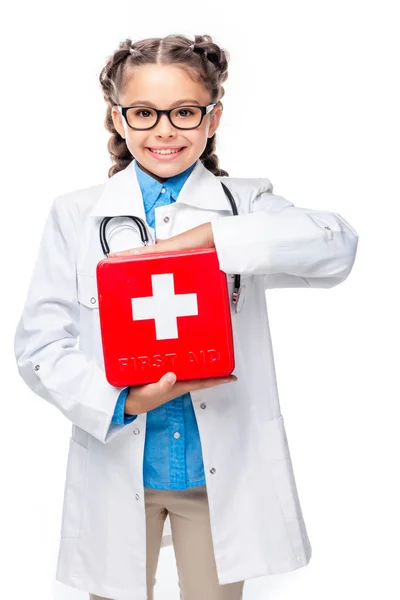 Sonriente escolar en traje de médico sosteniendo botiquín de primeros auxilios aislado en blanco - foto de stock