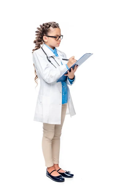 Visão lateral da criança em traje de médico escrevendo algo para prancheta isolada em branco — Fotografia de Stock