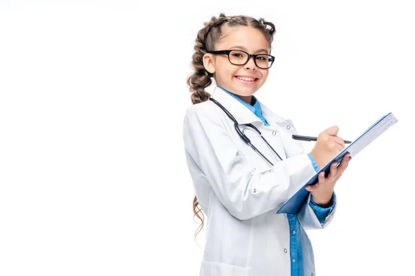 Sonriente escolar disfrazado de médico escribiendo algo al portapapeles aislado en blanco - foto de stock