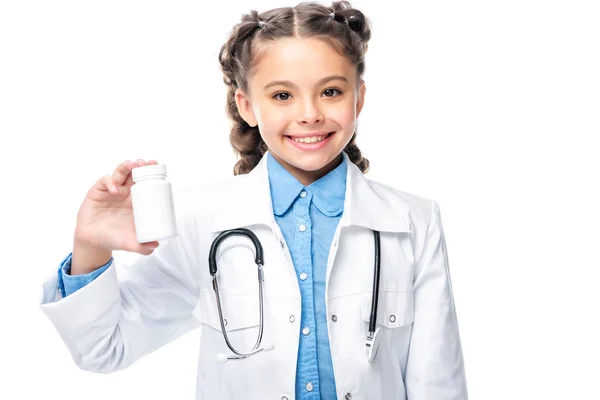 Sonriente escolar en traje de médico sosteniendo botella de píldoras aisladas en blanco - foto de stock