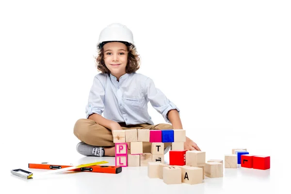 Schoolboy in costume of architect and helmet sitting near wooden cubes isolated on white — Stock Photo
