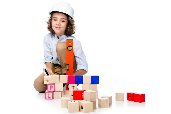 Schoolboy in costume of architect and helmet measuring wooden cubes with spirit level isolated on white — Stock Photo