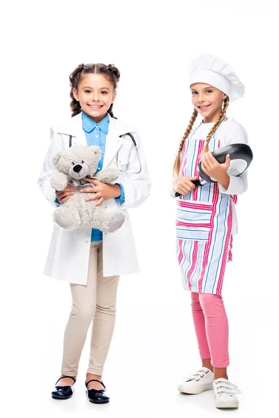 Schoolchildren in costumes of doctor and chef standing with teddy bear and frying pan isolated on white — Stock Photo
