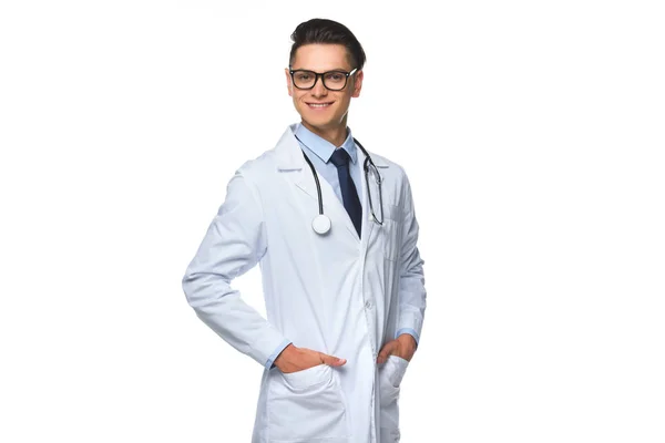 Glücklicher junger Arzt mit Händen in Arztkitteltaschen, isoliert auf weißem Grund — Stockfoto