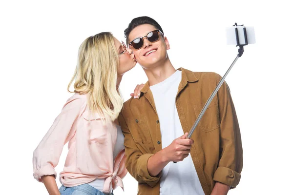 Jeune couple prenant selfie de monopode tandis que la femme embrasser son petit ami isolé sur blanc — Photo de stock