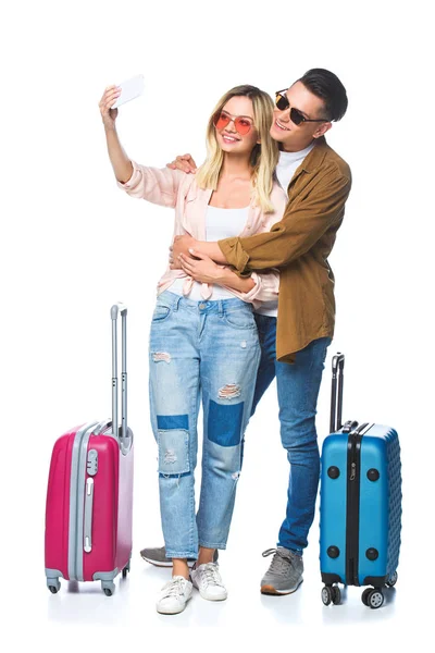 Молодая путешествующая пара с чемоданами, делающая селфи со смартфоном, изолированным на белом — стоковое фото