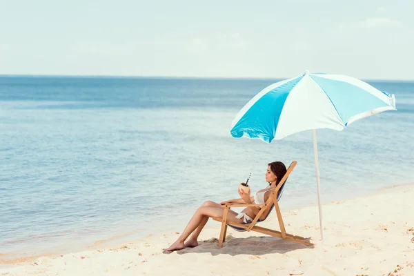 Beautiful girl in bikini with coconut cocktail resting on deck chair under beach umbrella near sea — Stock Photo