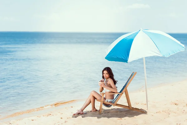 Mujer joven en bikini bebiendo coctel de coco en chaise longue de playa bajo sombrilla cerca del mar - foto de stock