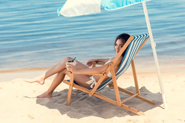 Attraktives Mädchen im Bikini mit Kokoscocktail am Strand unter Sonnenschirm — Stockfoto