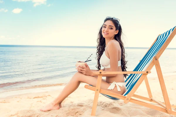 Jovem mulher de biquíni bebendo coquetel de coco enquanto relaxa na cadeira deck — Fotografia de Stock