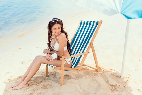 Счастливая женщина со смартфоном отдыхает на пляжном шезлонге под солнцем на морском курорте — стоковое фото