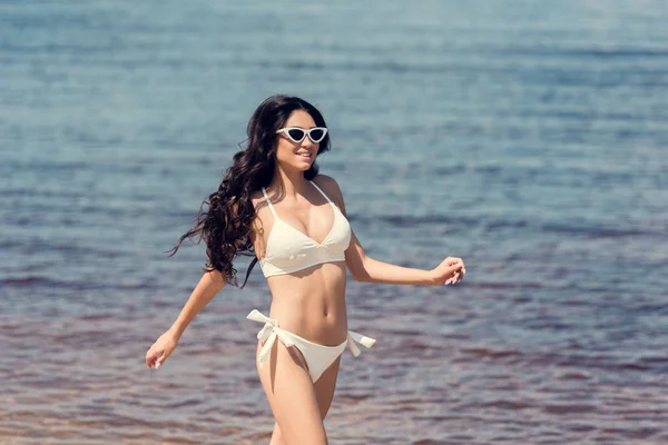 Happy woman in sunglasses and white bikini running near the sea — Stock Photo