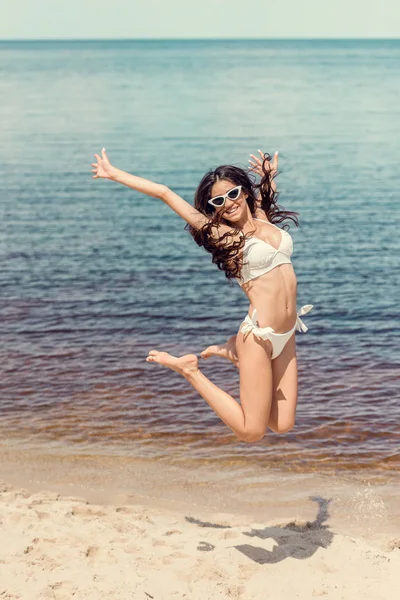 Mujer excitada en gafas de sol y bikini blanco saltando en la playa cerca del mar - foto de stock