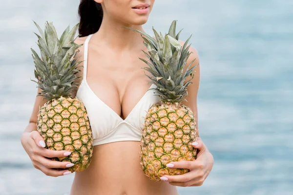Vista recortada de chica en bikini sosteniendo piñas frescas cerca del mar - foto de stock