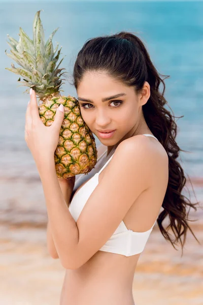 Portrait of young brunette woman in white bikini holding sweet pineapple near the sea — Stock Photo