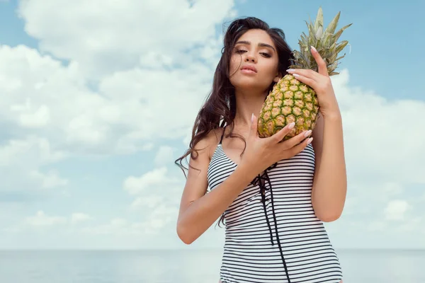 Attractive brunette girl in swimsuit posing with pineapple near the sea — Stock Photo