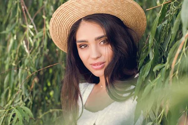 Portrait of attractive brunette girl in straw hat posing near willow tree — Stock Photo