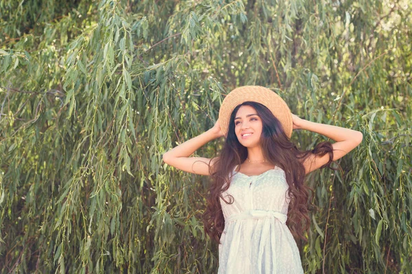 Усміхнена брюнетка дівчина в солом'яному капелюсі і біла сукня позує біля верби дерево — стокове фото