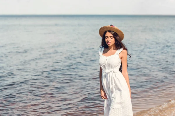 Mujer joven en sombrero de paja y vestido blanco caminando cerca del mar - foto de stock