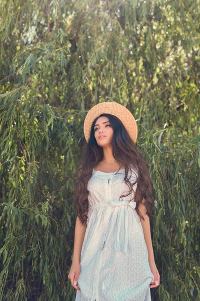 Beautiful girl in straw hat and white dress posing near willow tree — Stock Photo