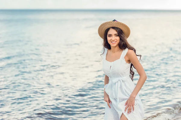 Mujer sonriente con sombrero de paja y vestido blanco caminando cerca del mar - foto de stock