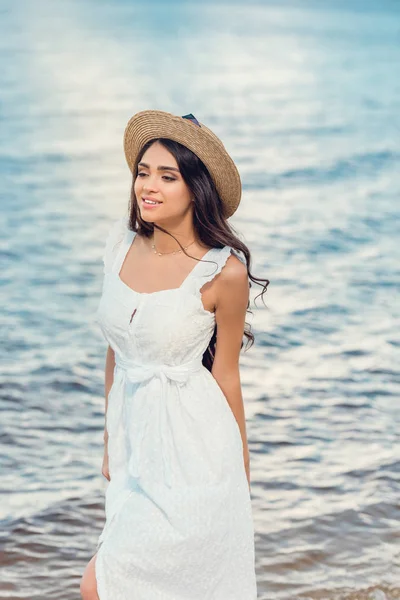 Schönes glückliches Mädchen mit Strohhut und weißem Kleid, das am Meer spaziert — Stockfoto