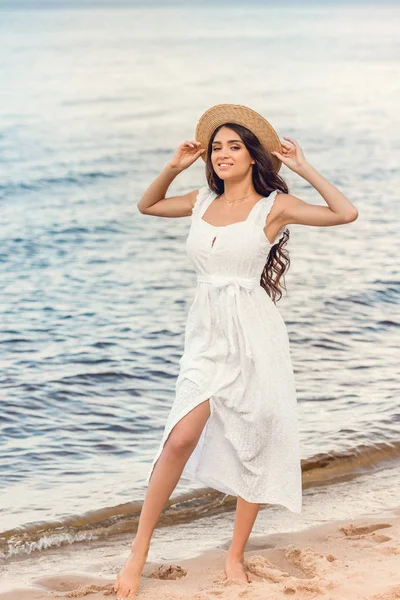 Attractive smiling woman in straw hat and white dress walking on seashore — Stock Photo