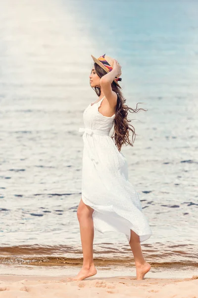 Jeune femme en chapeau de paille et robe blanche marchant sur la plage de sable — Photo de stock