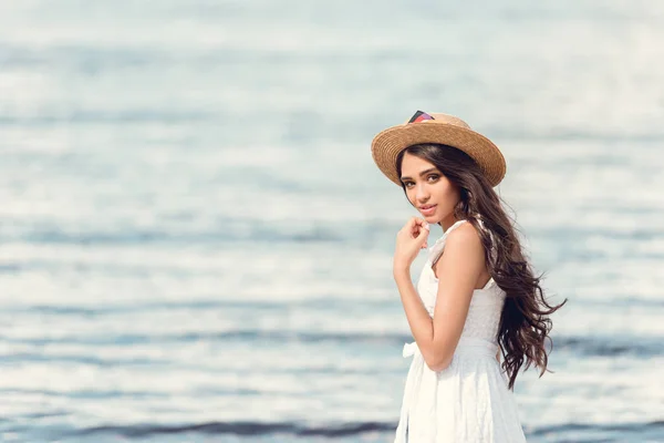 Attractive brunette girl in straw hat and white dress walking near the sea — Stock Photo