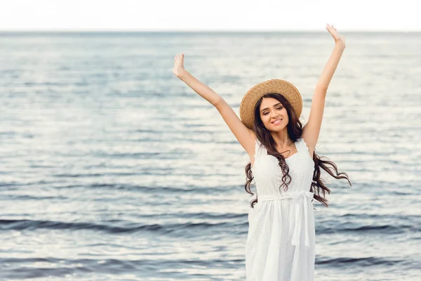Aufgeregtes brünettes Mädchen mit Strohhut und weißem Kleid am Meer im Sommer — Stockfoto
