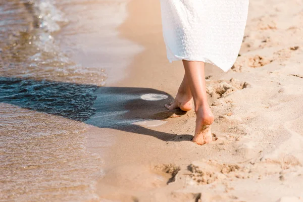 Vista parcial de chica descalza en vestido blanco caminando en la playa de arena cerca del agua - foto de stock