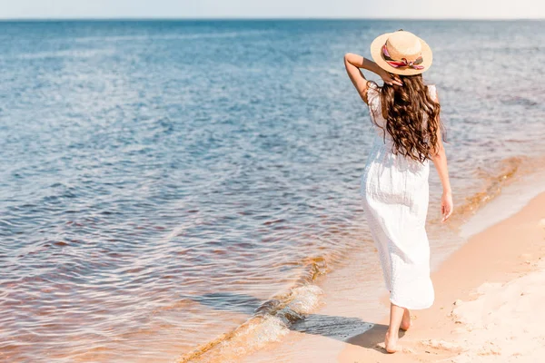 Back view of woman in straw hat and white dress walking on beach near sea — Stock Photo
