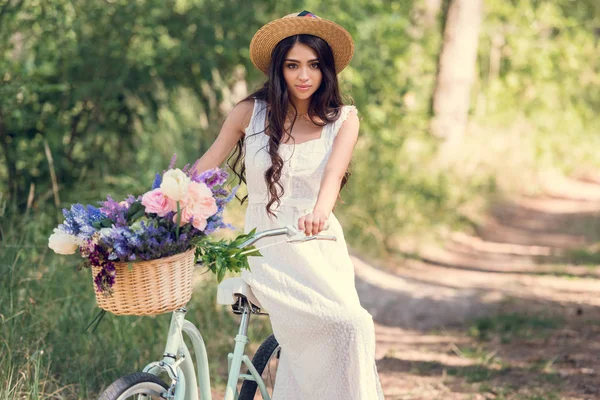 Chica atractiva en sombrero de paja sentado en bicicleta con flores en canasta de mimbre en el parque - foto de stock