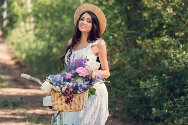 Hermosa chica en sombrero de paja y vestido blanco posando con bicicleta y flores en canasta de mimbre - foto de stock